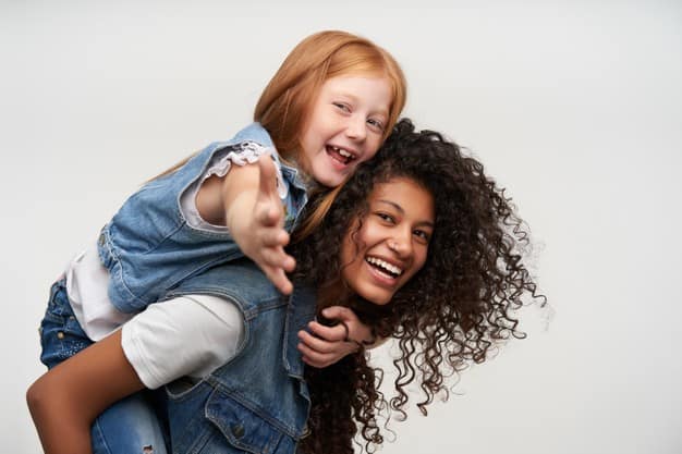 side-view-joyful-pretty-young-dark-skinned-brunette-woman-riding-her-back-cheerful-cute-redhead-female-kid-looking-happily-smiling-broadly-isolated-white_295783-7458